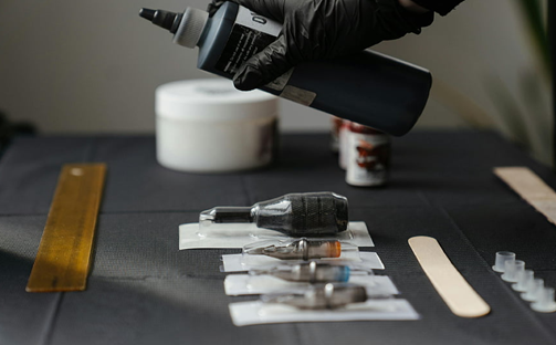Photograph of tattoo equipment ready for use within a tattoo studio, showing needles for tattoo gun in protective plastic, ink and ink pots, tub of petroleum jelly, emery board and ruler.